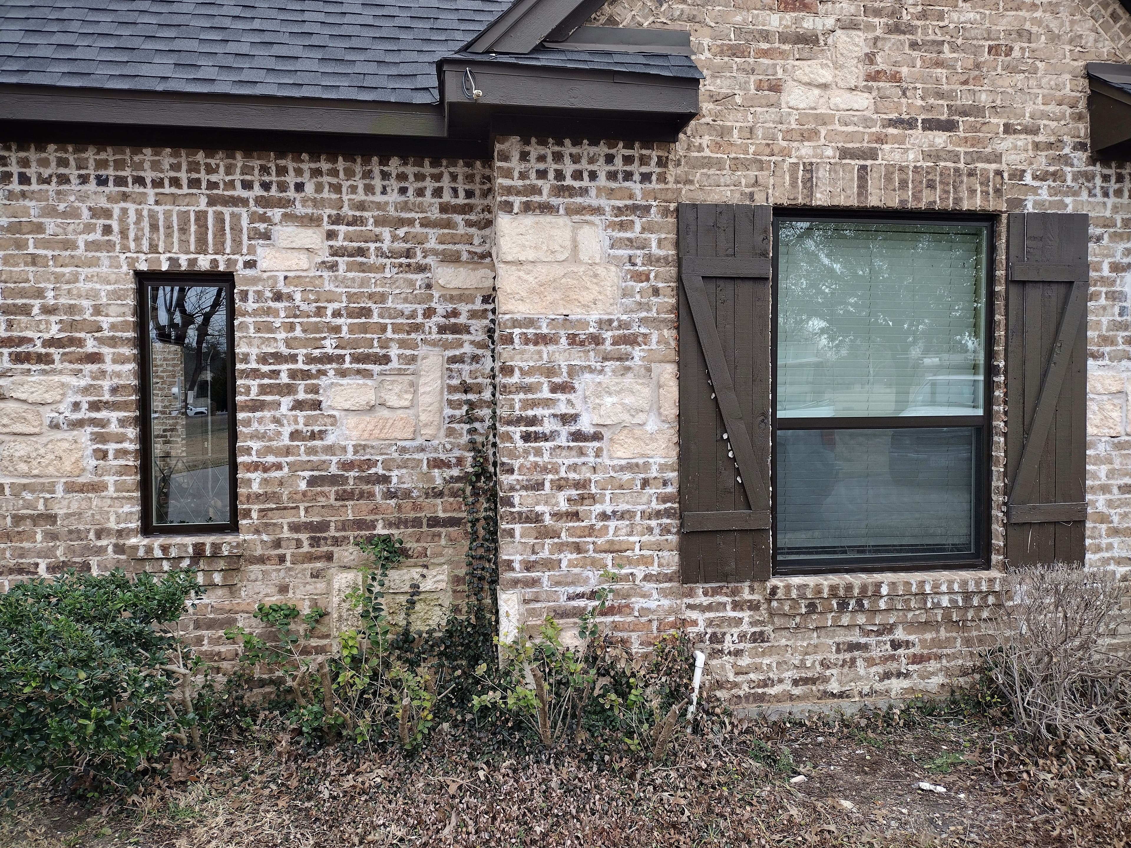 Black-framed replacement windows with rustic wooden shutters on tan brick and stone exterior